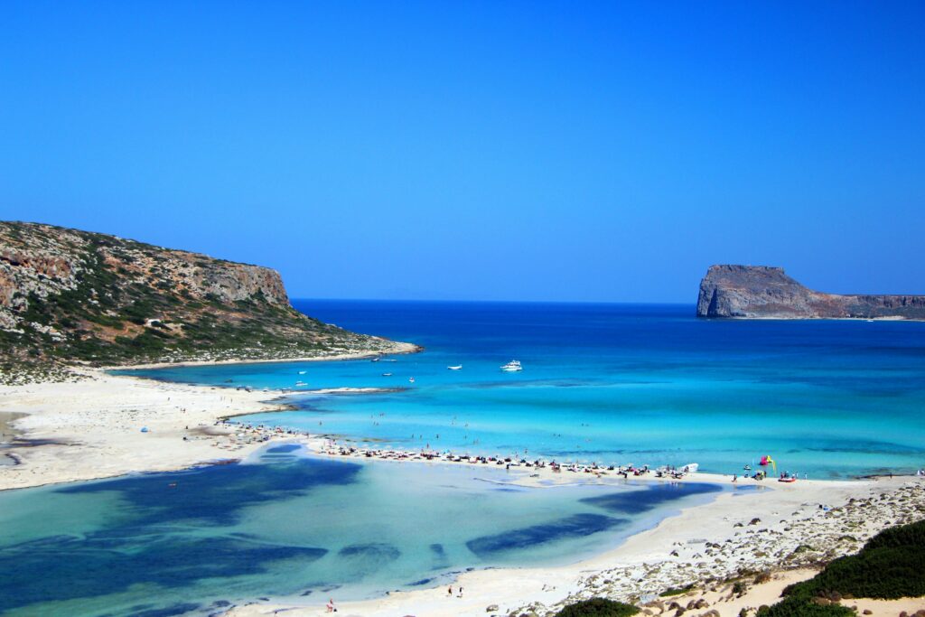 Secluded white-sand beach in Crete, framed by private cabanas with billowing linen drapes and turquoise waters reflecting a silver champagne tray.