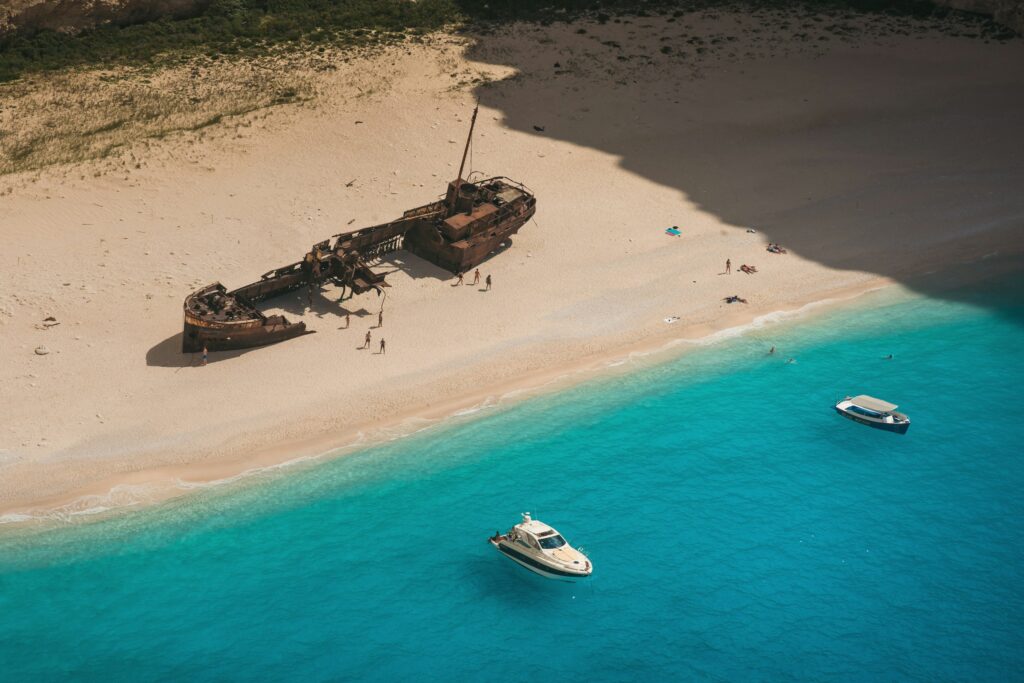 A boat approaches the iconic Shipwreck Beach in Zakynthos, Greece, with its rusted wreck framed by white sands and turquoise waters, while travelers explore the glowing blue chambers of the nearby sea caves under bright sunlight.