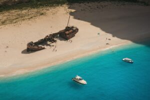 A boat approaches the iconic Shipwreck Beach in Zakynthos, Greece, with its rusted wreck framed by white sands and turquoise waters, while travelers explore the glowing blue chambers of the nearby sea caves under bright sunlight.